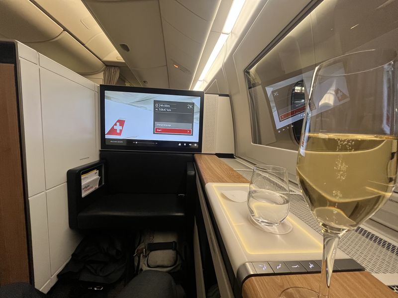 Business class seat on a Swiss aircraft, the screen showing 24 hours and 10,647 km to go, a glass of white wine on the table.