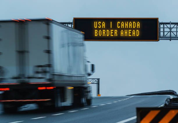 Highway sign reading USA Canada Border Ahead, a truck passing beneath it at dusk.