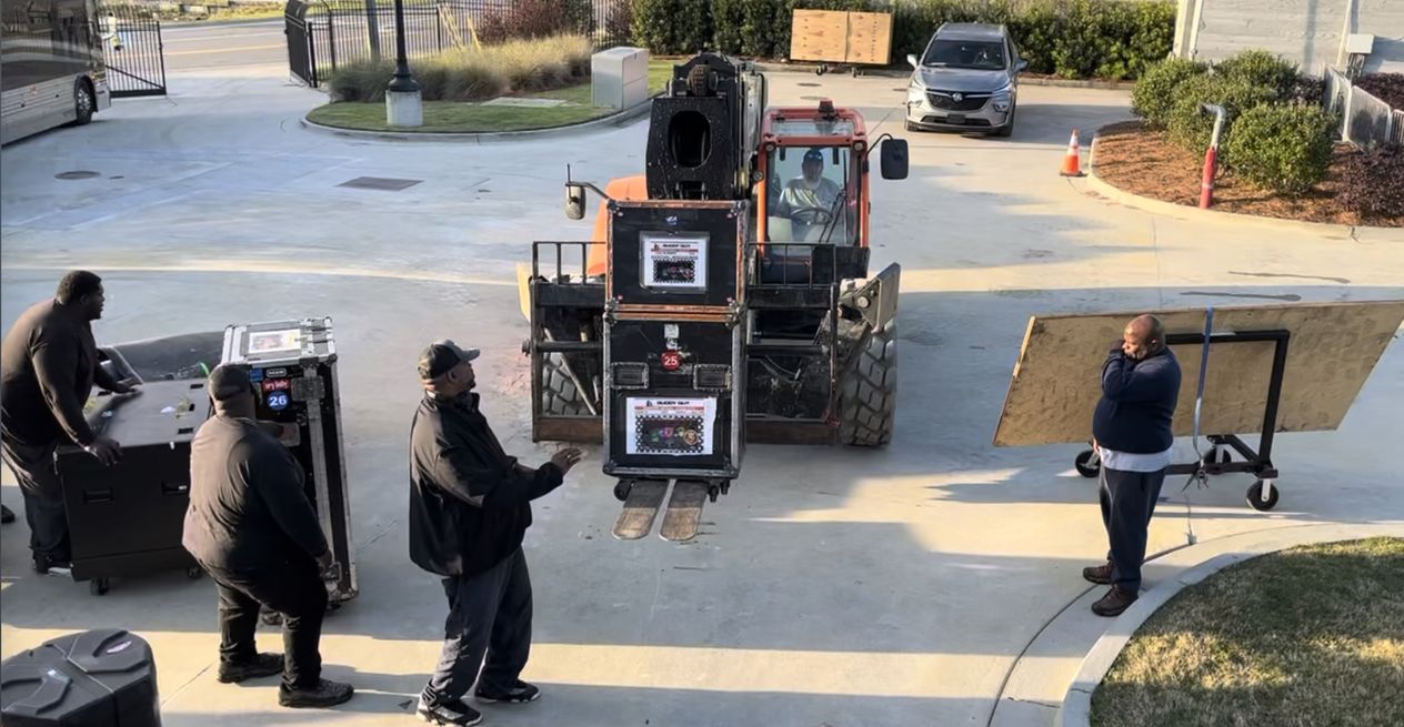 Crew watching a telehandler carry stacked road cases across a venue forecourt.