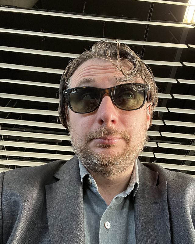 Sage Anthony in sunglasses and a grey jacket, venue seating behind him.