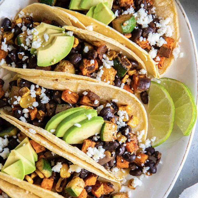 Three tacos on a plate with avocado, black beans, crumbled cheese and a wedge of lime.