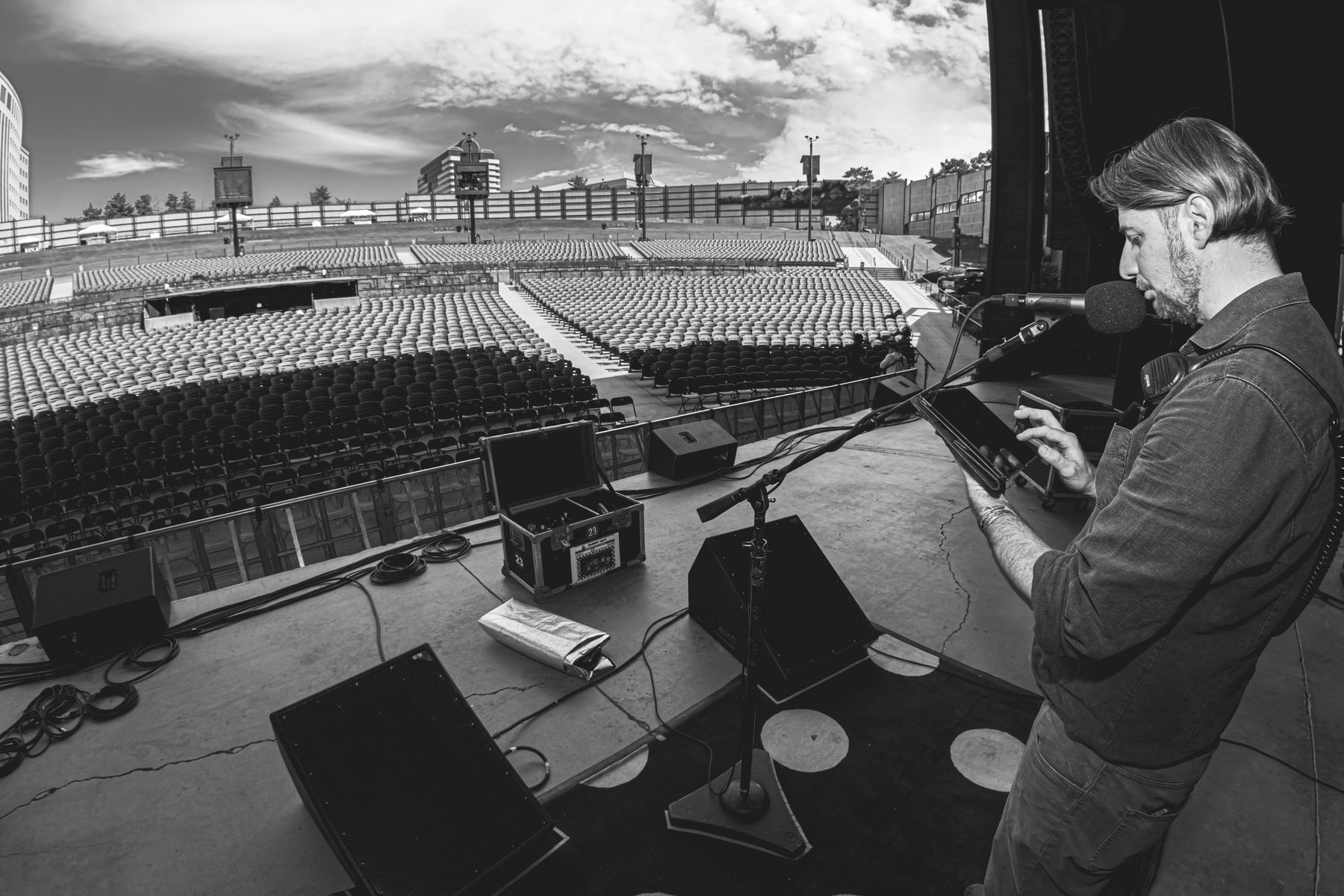 Black and white shot of an engineer on stage with a tablet, talking into a talkback mic, empty amphitheatre seats stretching back.
