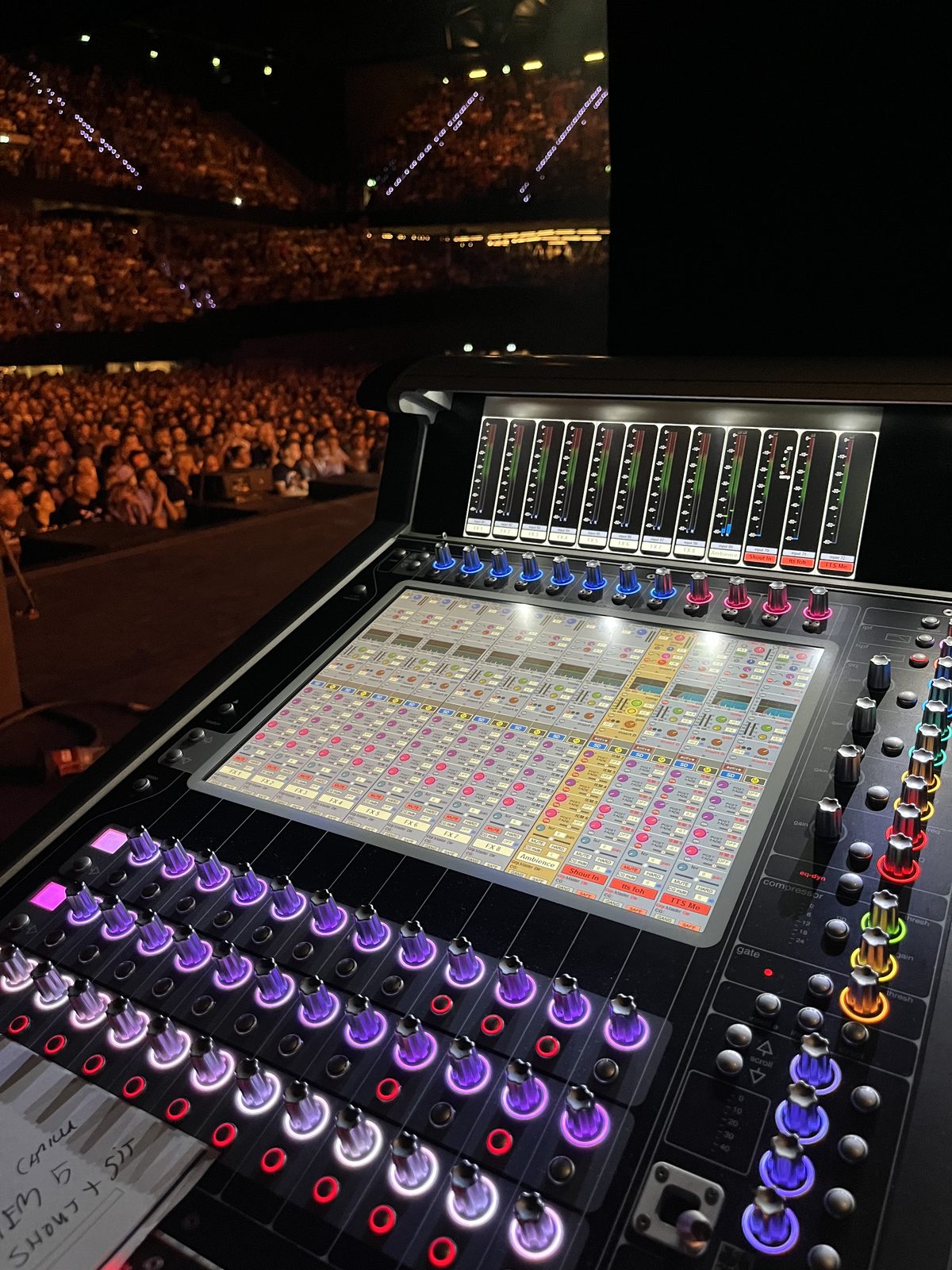 Digital mixing console lit up during a show, channel names on the screen, arena crowd out beyond the desk.