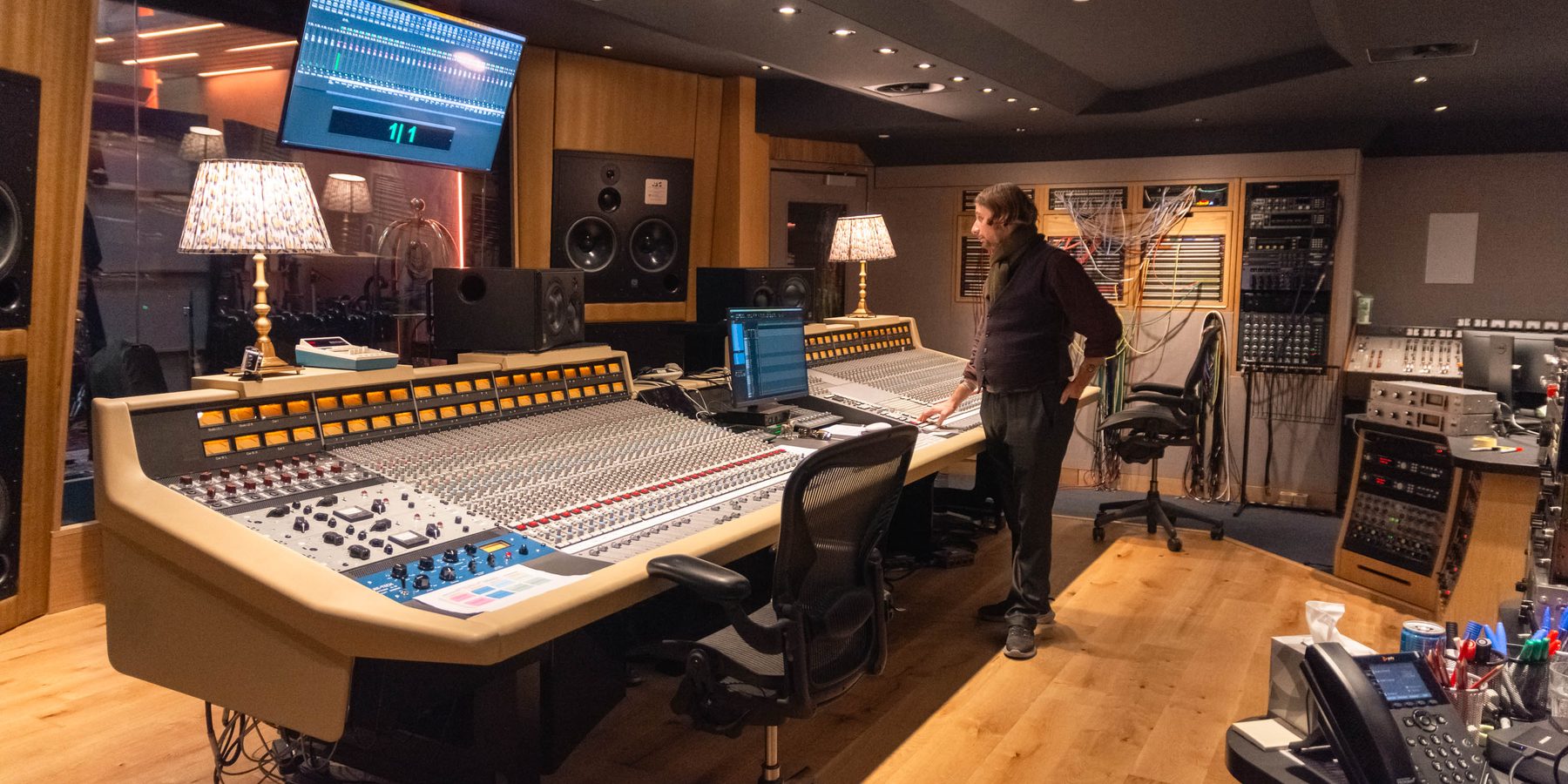 Studio control room, engineer standing at a large analogue console, main monitors soffit mounted, outboard racks and patchbay to the right.