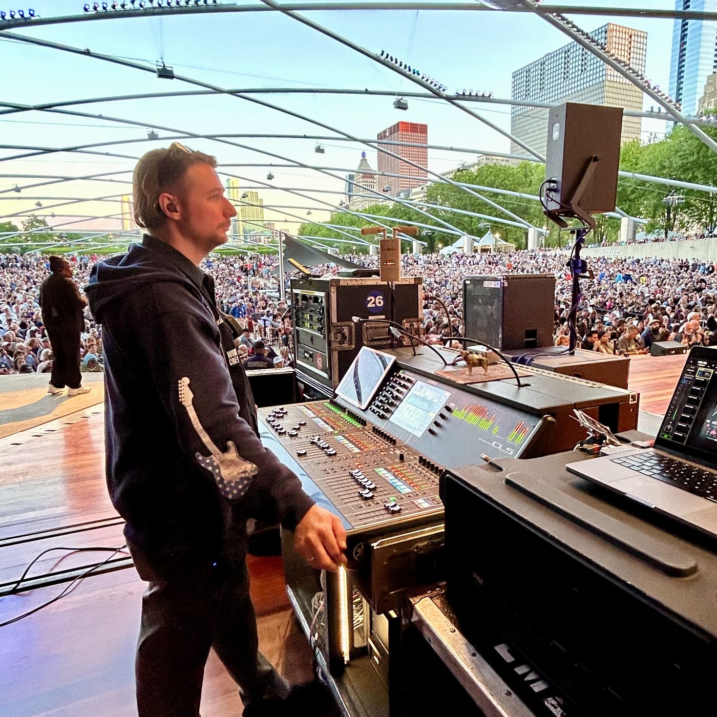 Engineer at a Yamaha CL5 beside the stage, crowd filling the lawn under a steel trellis with a city skyline behind.