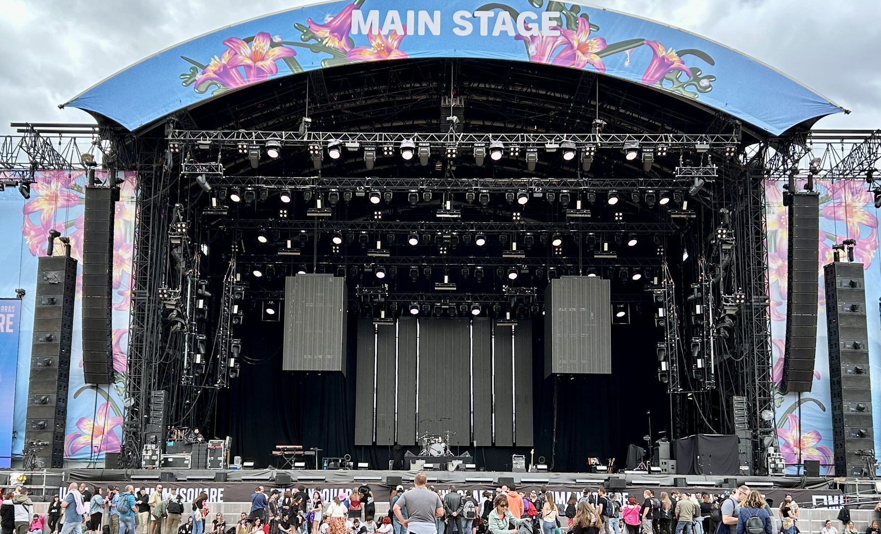 Empty festival main stage before doors, line arrays flown and stacked, lighting rig hung, drum kit alone at centre.