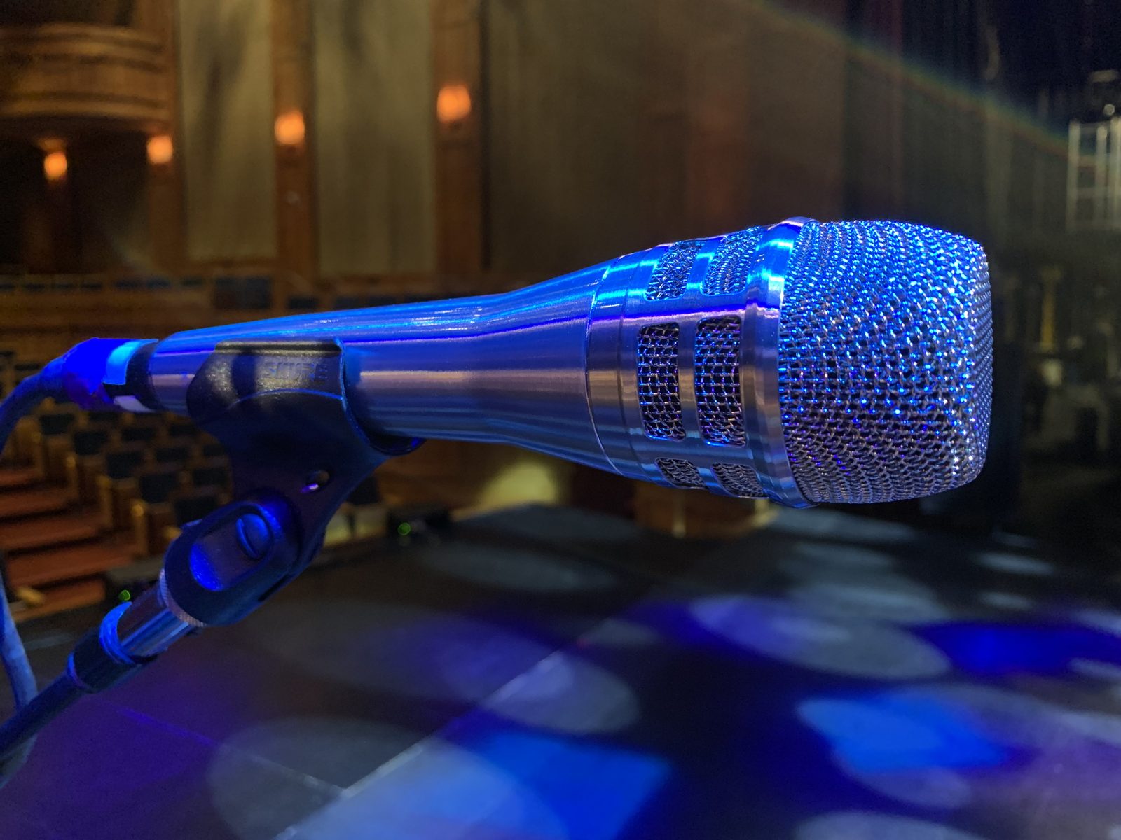Close up of a Shure vocal microphone on its stand under blue light, empty theatre seats behind.