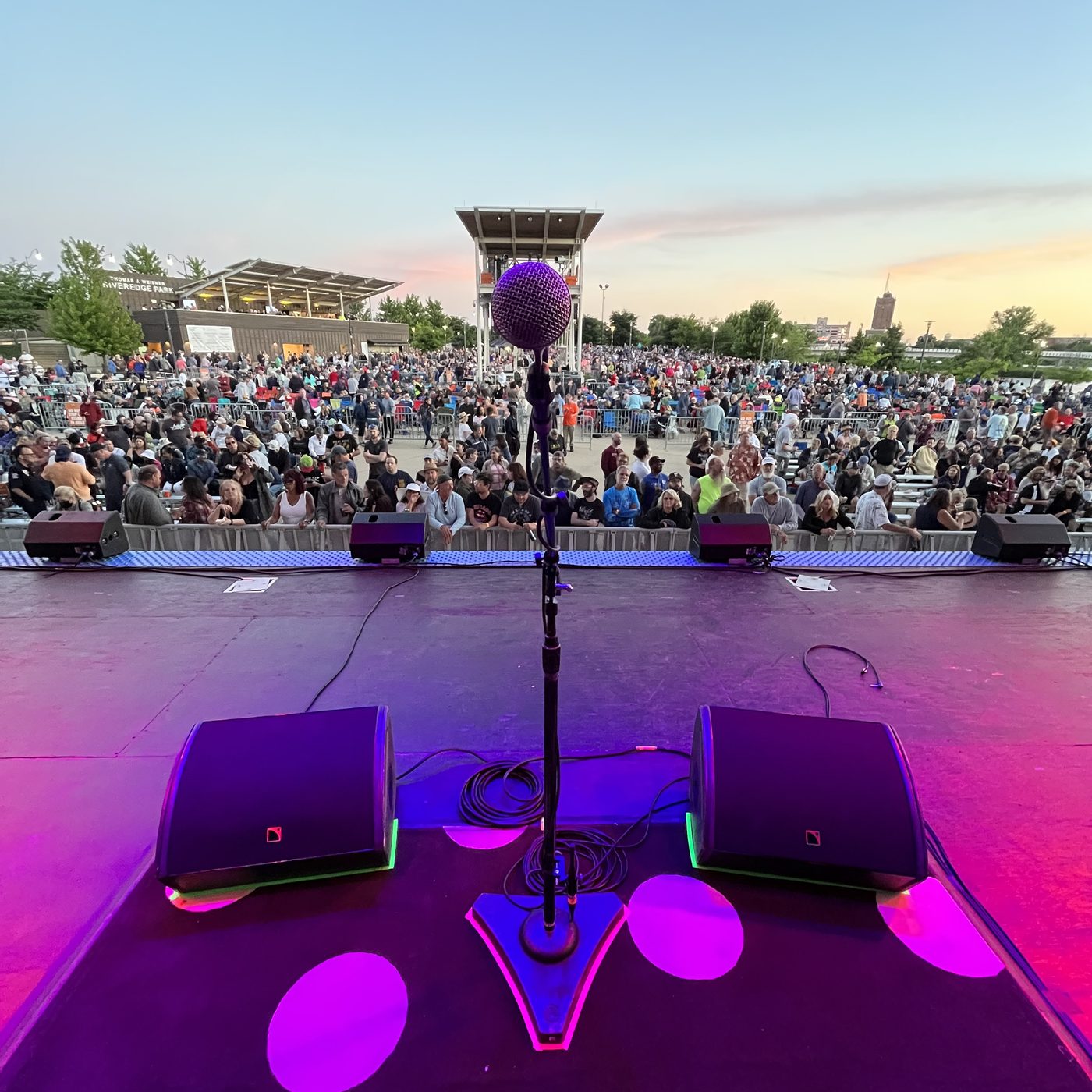View from behind the vocal mic at dusk, two L-Acoustics wedges either side, crowd filling the park beyond the barricade.